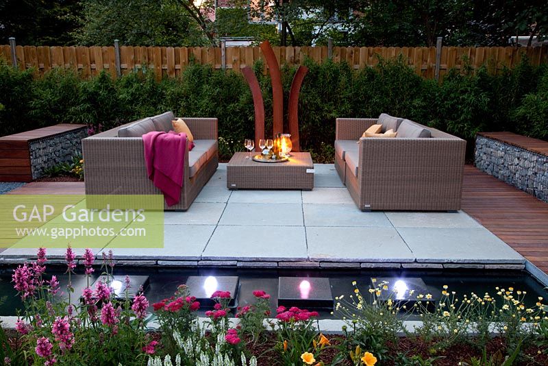 Small garden lit up at night, with wicker sofas on decked and paved patio, backed by Fargesia murielae - Bamboo hedge. Rectangular pond with row of square water features and lights  