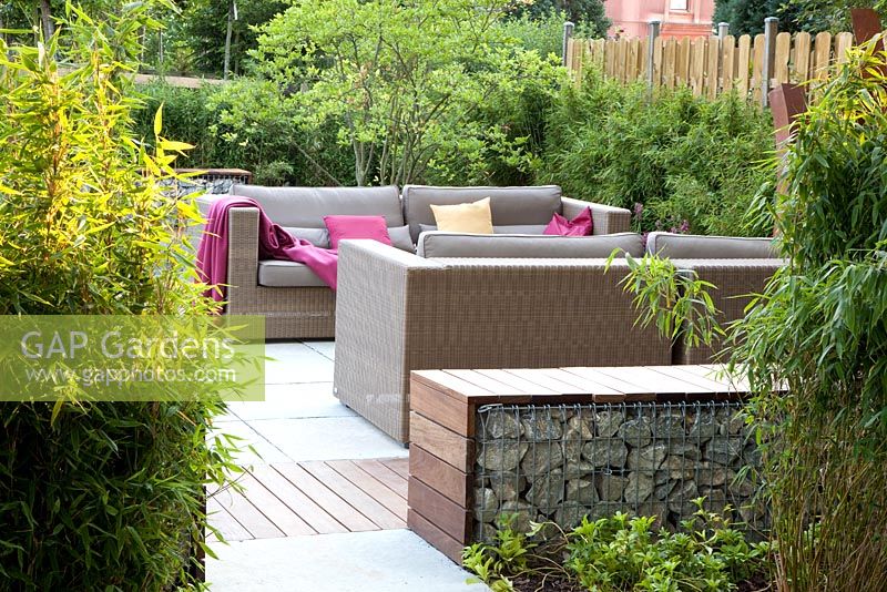 Small garden with wicker sofas and Gabion benches on decked and paved patio, backed by Fargesia murielae - Bamboo hedges.