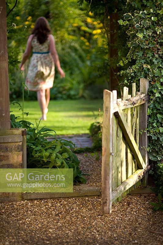 Woman wearing a summer dress, walking through a rustic wooden garden gate to the lawn beyond