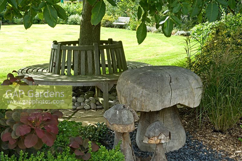 Circular bench under tree and wooden ornamental mushrooms. Parsons Cottage, Essex, UK