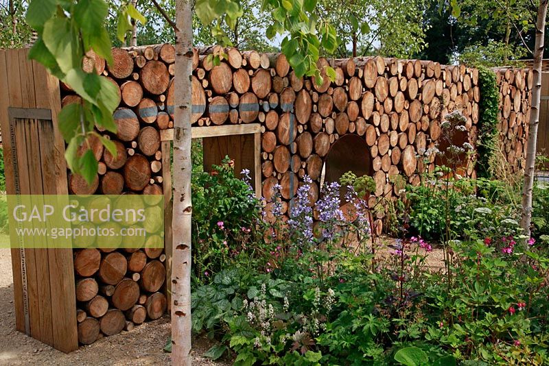 Narratives of Nature garden with log wall and moon gate. Planting includes ferns, Geranium sylvaticum, Luzula nivea, Atriplex hortensis, Umbilicus rupestris and Betula pubescens - The Future Gardens, St Albans, Herts 