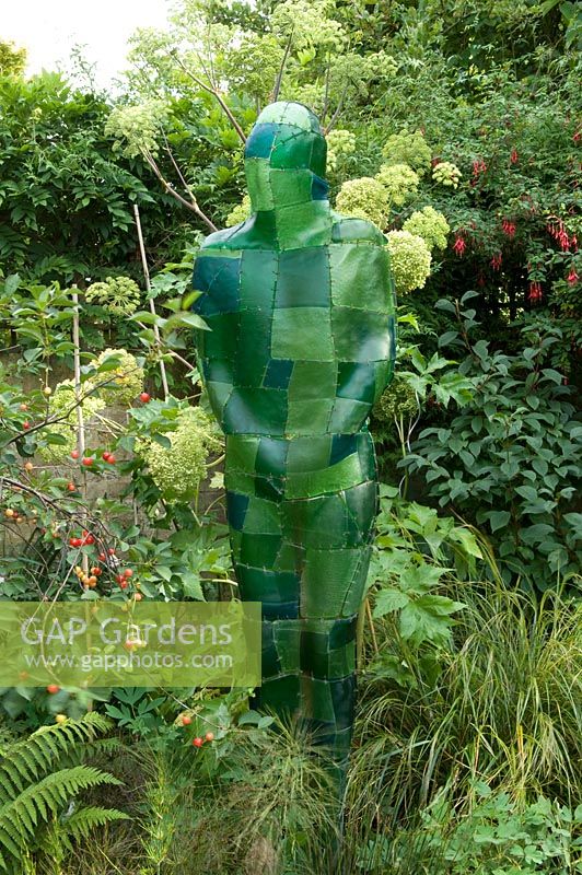 Green resin sculpture figure in border with Angelica and Prunus - Cherry tree. Yulia Badian garden, London, UK
 