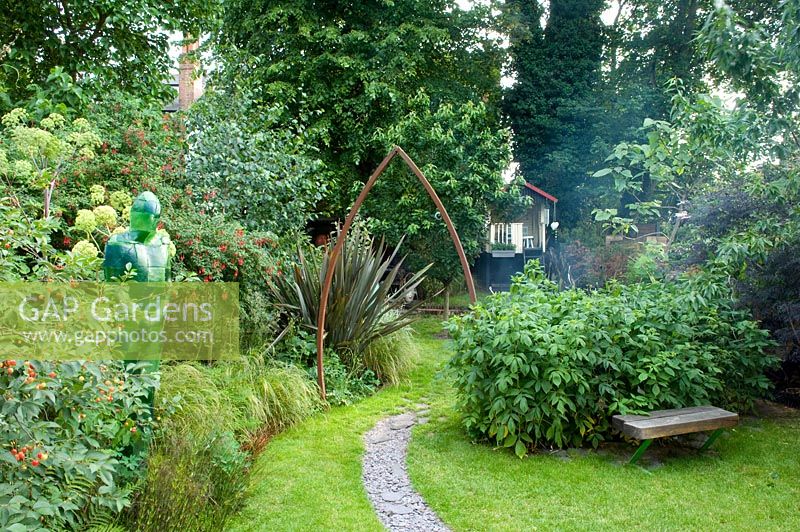 Urban garden with metal arch over slate path. Resin statue in border with Phormium -New Zealand Flax. Wooden bench on lawn. Yulia Badian, London, UK 
 