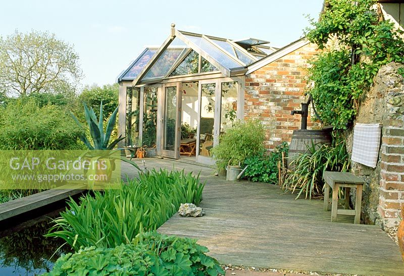Timber decking, pond and conservatory. Wooden bench and Agave in container. Late Spring, Fovant Hut Garden, Wilts