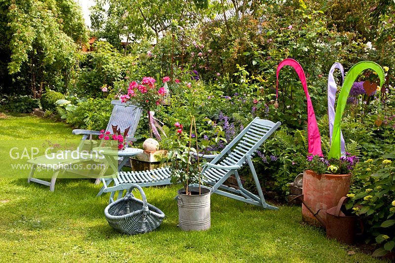 Loungers on lawn next to border with Rosa and colourful ornament made from strips of fabric in terracotta pot
 
