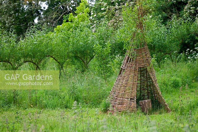 Woven willow tepee with tree trunk seat, in front of living Willow arched hurdles