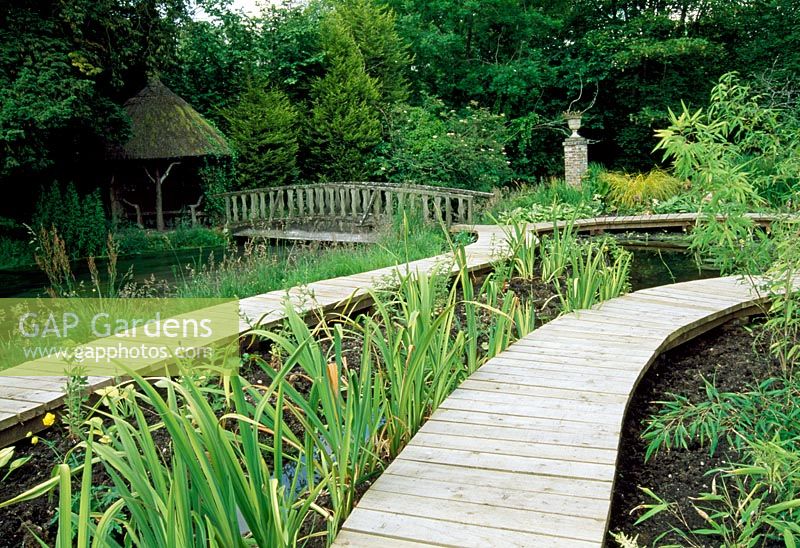 Raised boardwalk winding through bog garden, bridge over river leading to summerhouse - Weir House, Hants