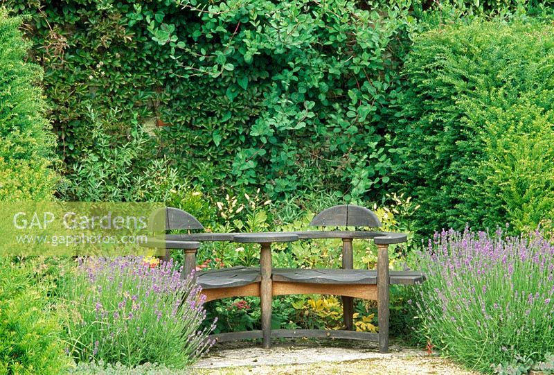 Wooden seat, designer Matthew Burt in border with Lavandula, Lilium and topiary yew - Weir House, Hants
