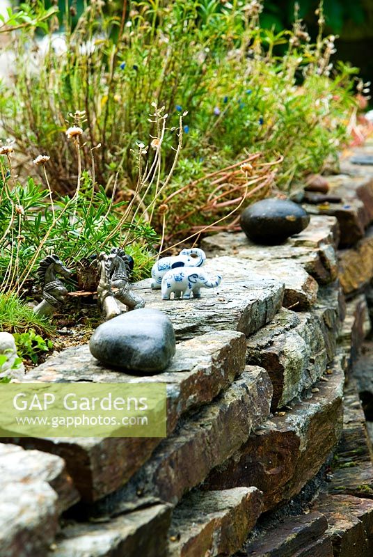 Stone wall planted with Sedums and alpines and decorated with small figurines and statuettes for the benefit of younger visitors - Pinsla Garden, Cardinham, Cornwall