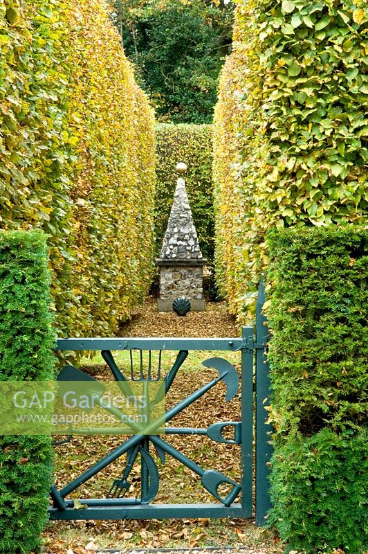 Blue gate leading to clipped Fagus and Taxus baccata hedges and stone obelisk - Silverstone Farm, Norfolk
