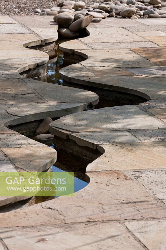 A meandering canal leads through a flagstone paving towards a beach with pebbles that belongs to a natural swimming pool. 