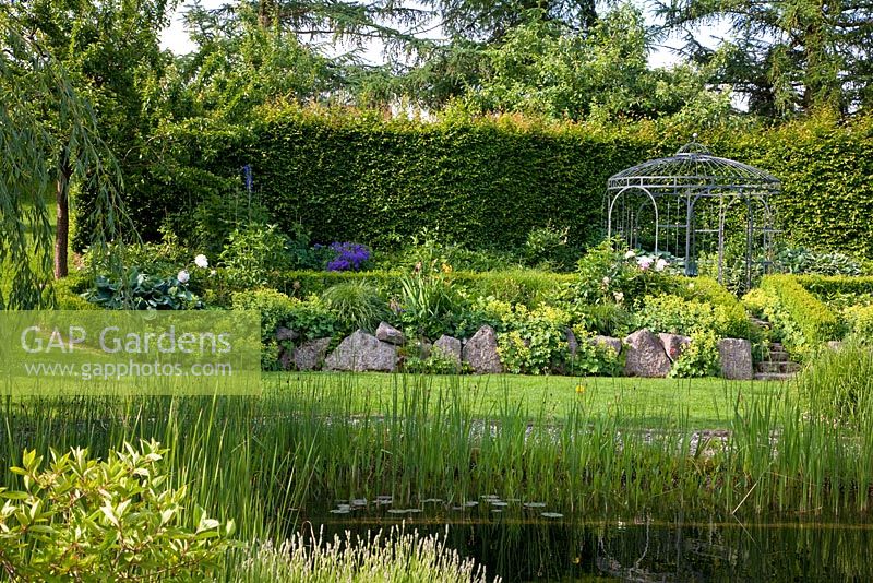 View over a natural swimming pool to a pavillon, backed with a hedge. The slope is secured by lime sand boulders, planting includes Alchemilla mollis, Buxus, Carpinus betulus, Fagus, Geranium, Hosta, Larix, Lupinus and Salix
