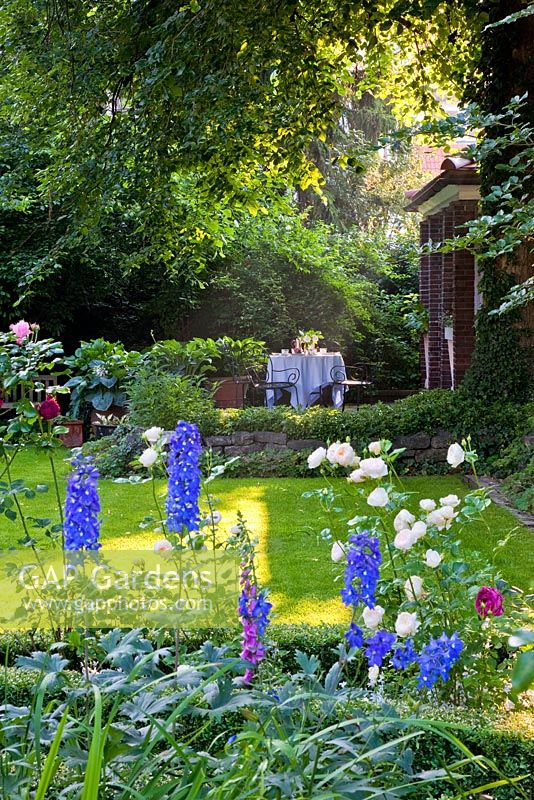 Sheltered by a beech tree, a table and chairs on the raised veranda of a brick pavilion viewed through mixed border of Delphinium Elatum-Grp and Fagus sylvatica. The Manor House, Germany 
