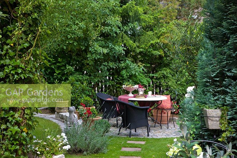 Seating area with a decorated table, black furniture and containers on paved circle. Other planting includes Chamaecyparis lawsoniana, Hedera helix and Lavandula