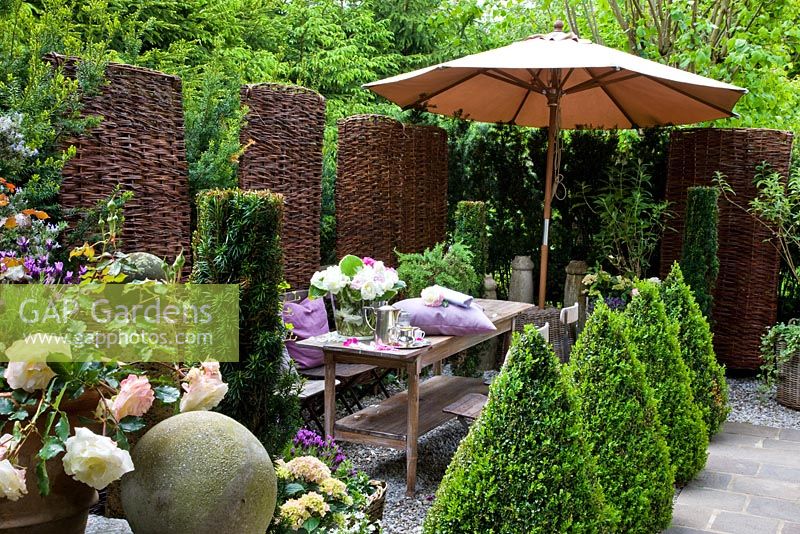 A patio with a wooden table and a parasol, flanked by a row of box pyramids and wicker trellis