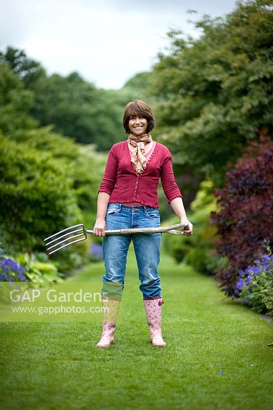 Woman wearing blue jeans and wellies standing on a lawned path carrying a garden fork 