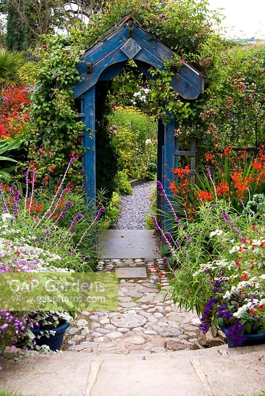 Wooden archway surrounded by a mass of colourful, flowering plants including Crocosmias, Salvia leucantha, Helichrysum and Clematis. Poppy Cottage Garden, Roseland Peninsula, Cornwall, UK