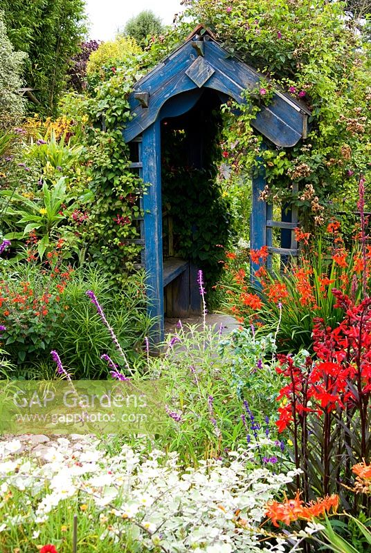 Wooden archway surrounded by a mass of colourful, flowering plants including Crocosmias, Salvia leucantha, Lobelia cardinalis 'Queen Victoria', Helichrysum and Clematis. Poppy Cottage Garden, Roseland Peninsula, Cornwall, UK