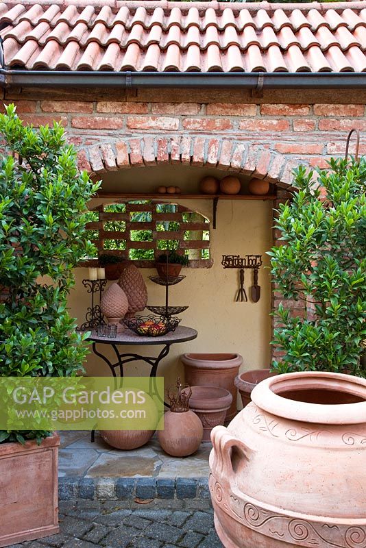 Patio and gangway with bistro table, terracotta urns, objects and containers planted with Laurus nobilis