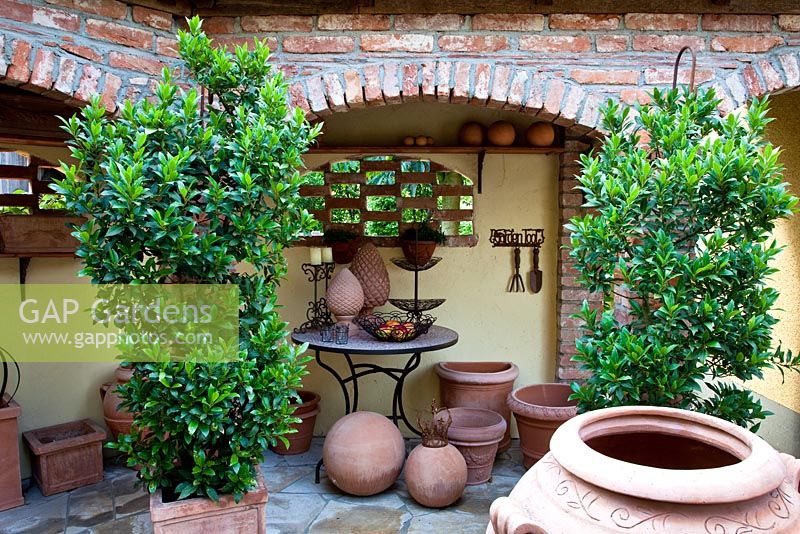 Display of terracotta and decorative metal objects on a bistro table in a gangway. Plants in containers are Laurus nobilis