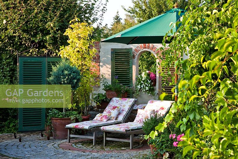 Next to backdrop with green shutters two loungers beneath green parasol on a granite paved rest area surrounded by Juniperus 