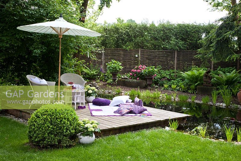 White wicker garden furniture, a blanket with cushions and a parasol on a wooden deck next to a garden pond. Willow trellis and hedge in the background. Plants are clipped Buxus, Fern and Hydrangea