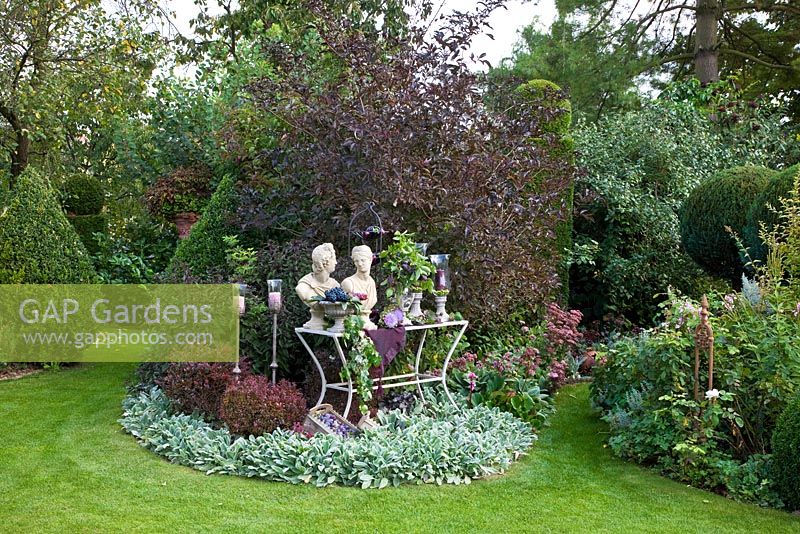 Etagère table with torsos and candle holders in a border with grey and purple leaved plants - Berberis thunbergii 'Atropurpurea Nana', Buxus topiary, Geranium macrorrhizum 'Ingwersen', Pinus, Sambucus nigra 'Black Beauty', Stachys byzantina 'Big Ear' and Stachys byzantina 'Silver Carpet'