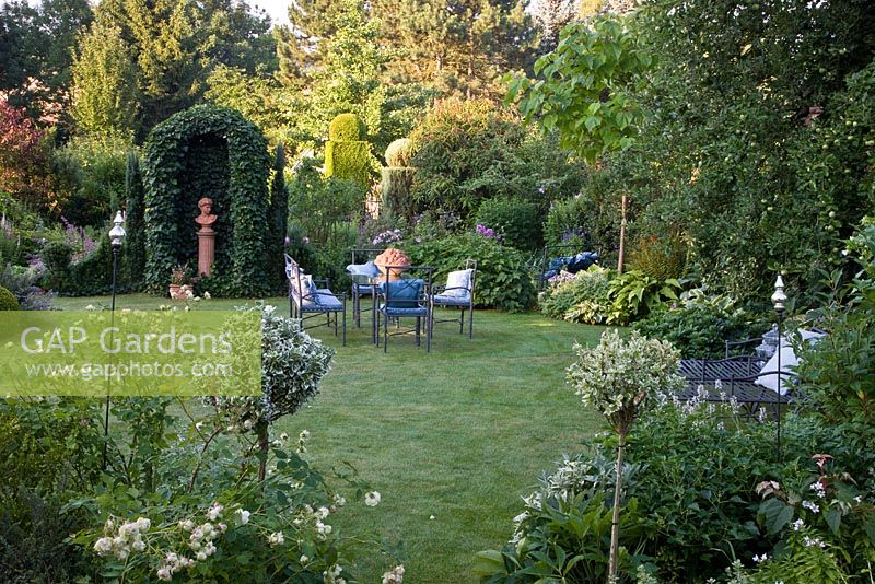 Country garden with metal garden furniture on lawn, clay torso on a plinth under Hedera helix 'Arborescens' - Ivy arch.  Borders of Euonymus fortunei 'Silver Queen', Hosta, Taxus baccata 'Fastigiata', Thuja occidentalis 'Smaragd'