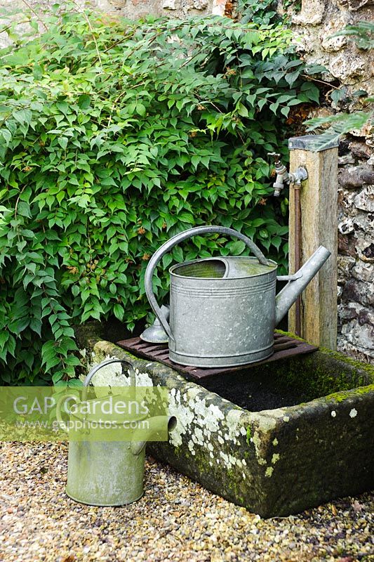 Watering cans beside outdoor tap mounted over old stone sink - Barnwells, Cerne Abbas, Dorset.