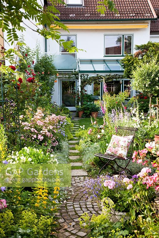 Stepping stones lead from the house to a round paved rest area with metal chair in a small garden with Rose 'Ballerina', Digitalis purpurea, Lobelia erinus, Lysimachia punctata, Nepeta Faassenii-Sorte and Petunia Surfinia