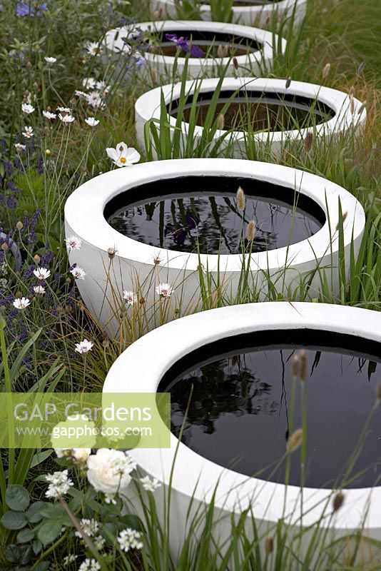 Circular white vessels containing dark pools of water set within meadow-style planting - Celebration and Jubilation - RHS Hampton Court Flower Show 2012
