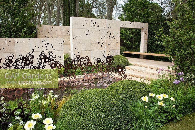 A garden inspired by the Arts And Crafts Movement. Formal paths and terraces combine with a water channel. Monolithic stone walls and an oak bench that appears to float. Focal point is an energy wave sculpture made from copper rings. Planting style is woodland edge with buxus domes - The M & G Garden