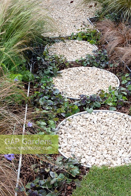 Path of circular containers filled with gravel, bark chippings and Viola with edging by ornamental grasses in the 'One Man Went To Mow' garden,  RHS Tatton Flower Show 2012