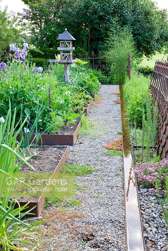 Modern kitchen garden with corten steel framed borders, a water rill, gravelled path and a wooden bird feeding house. Plants include Saponaria ocymoides