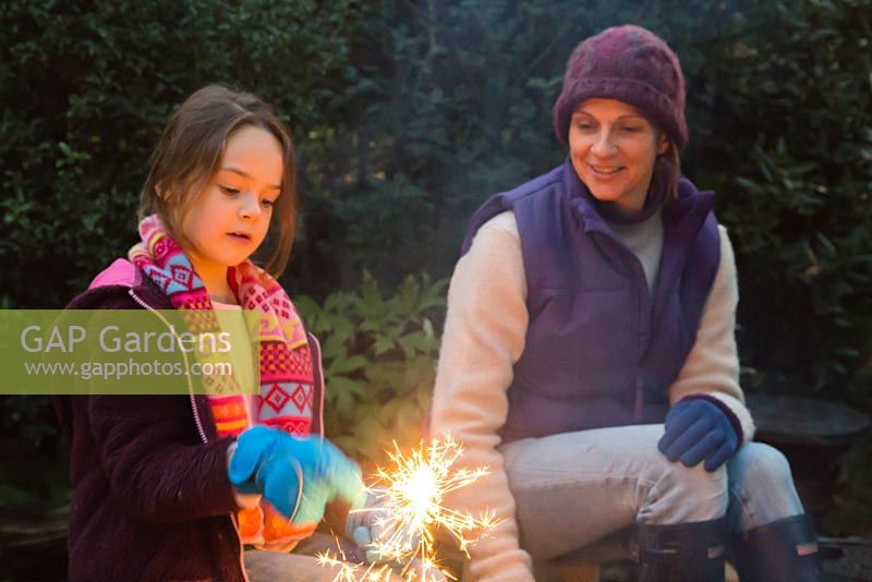 Mother and daughter lighting sparklers by a firepit in an autumnal back garden.