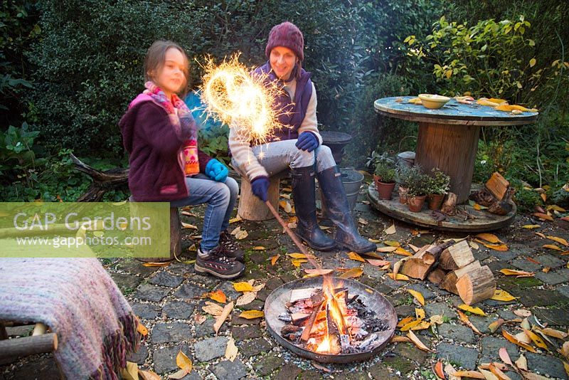 Mother and daughter lighting sparklers by a firepit in an autumnal back garden.