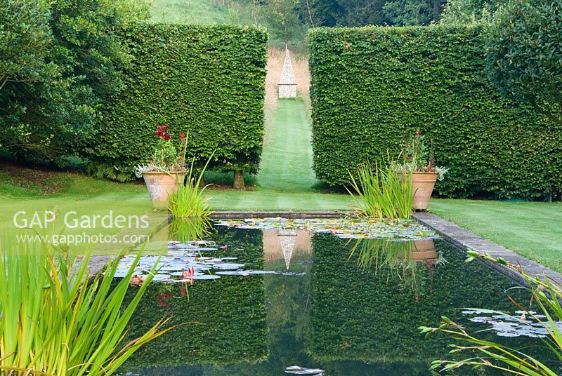 Formal pond lined by standard Crataegus x lavallei in the upper garden where a gap between beech hedges frames an obelisk set in a grassy meadow. Corscombe House, Corscombe, Dorset, UK