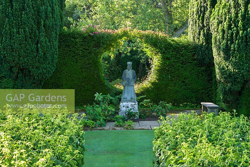 Statue of oriental priest is framed by a circle cut into a yew hedge that supports a honeysuckle, viewed from top of steps leading through wall clothed in Hydrangea petiolaris. Melplash Court, Bridport, Dorset, UK