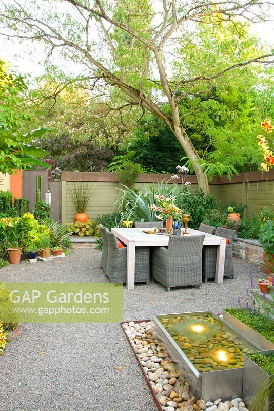 Summer outdoor living space with dining table and chairs lighted fountain, and enclosed with wooden fence. Albizia julibrissin - Persian Silk Tree syn. Mimosa Tree.