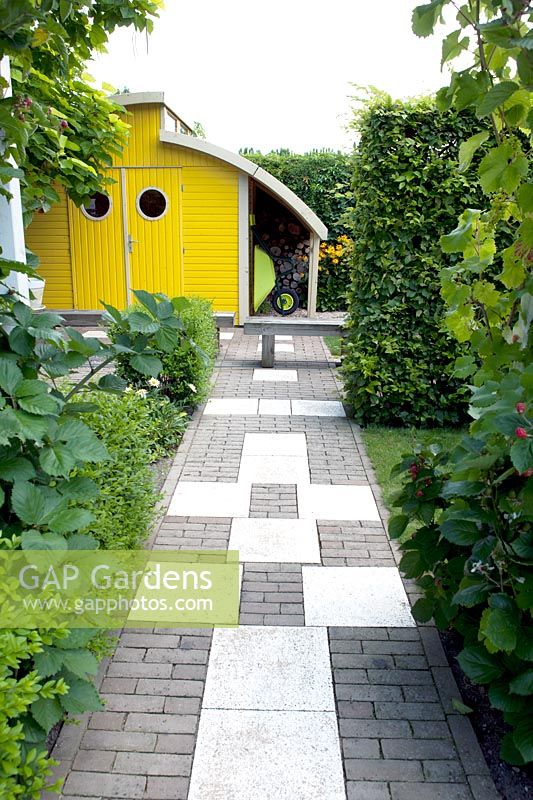Black and white pavement in the entrance. Bright yellow shed with roundshaped windows. Rudbeckia fulgida Goldsturm in the border.
