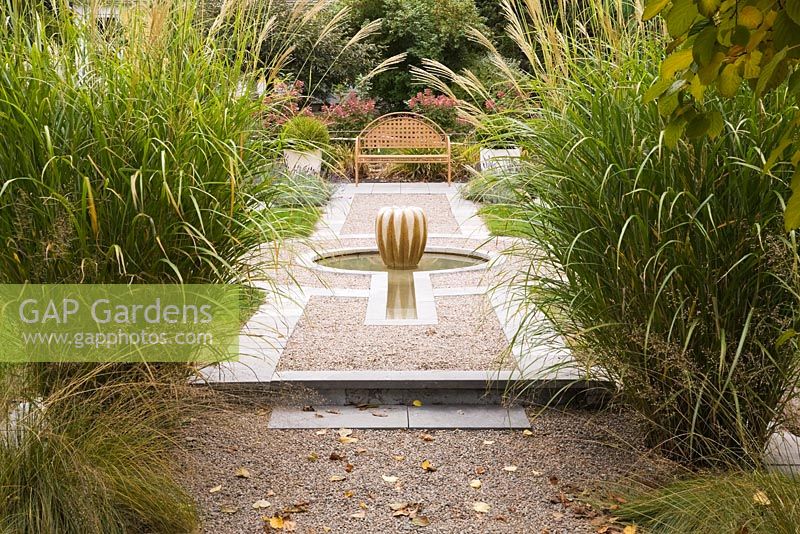 Raised gravel bed with gourd-shaped water fountain and brown metal lattice garden bench with pink hydrangeas paniculata 'Quick Fire' framed by tall (Miscanthus sinensis 'Berlin', ornamental grasses in backyard garden in autumn. Il Etait Une Fois garden, Monteregie, Quebec, Canada. 