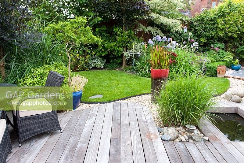 Hardwood Ipe deck with rattan furniture, formal pond, Miscanthus sinensis, ceramic pot with Imperata cylindrica 'Rubra' with lawn and herbacious border beyond 