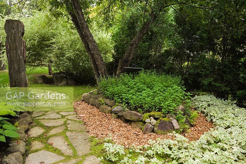 Jack-rabbit Johanssen wooden totem pole and flagstone path next to a raised border with Hostas and a Malus sylvestris - Wild Apple tree underplanted with Mentha - Mint, Melissa officinalis - Lemon Balm and Aegopodium podagraria - Goutweed 