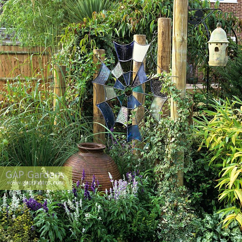 Stained glass spiders web suspended from ivy clad wooden posts. Blue salvia spires around huge pot below handcrafted, 