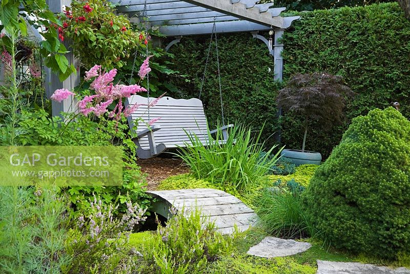 Pink Astilbes next to a footbridge and gray painted wooden swing bench underneath a pergola decorated with red Fuchsia flowers in a hanging basket in backyard garden in summer, Jardin Secret garden, Quebec, Canada