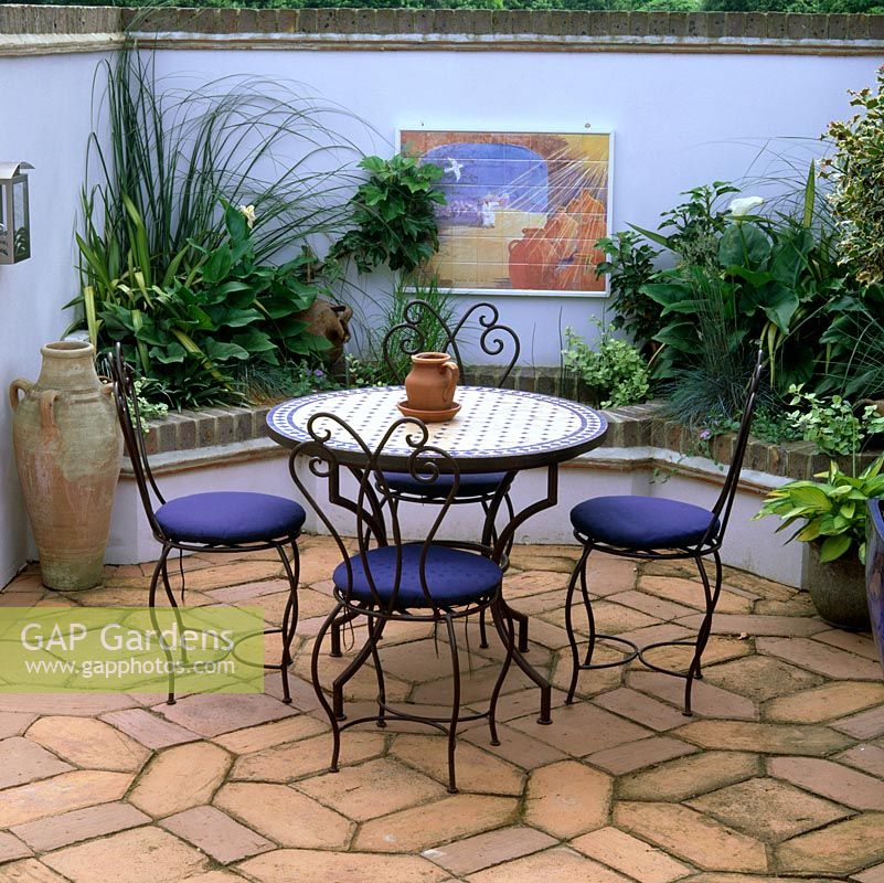 Moroccan style courtyard. Terracotta tiles, wire chairs and tiled table. Mosaic on wall above pool fed by gushing pitcher. Raised beds of arum, phormium, fatsia and grasses.