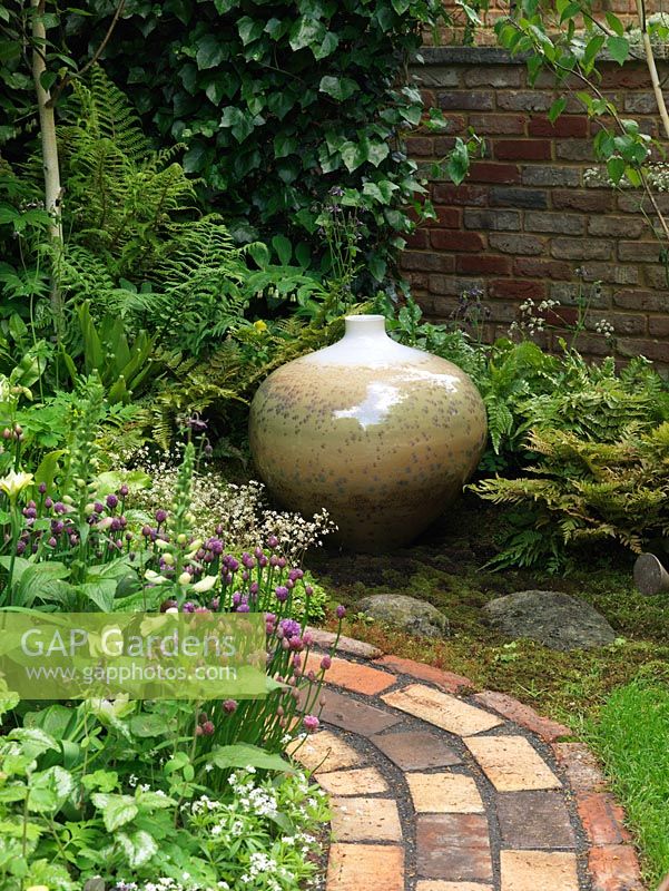Brick path circles bed of chives, foxglove and saxifrage. In shady corner surrounded by ferns, a lovely pot with crystalline glaze by potter John Stroomer.