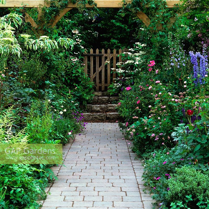 In front garden, wood gate opens onto stone path edged in roses, delphinium, hardy geranium, cistus, salvia and astrantia.