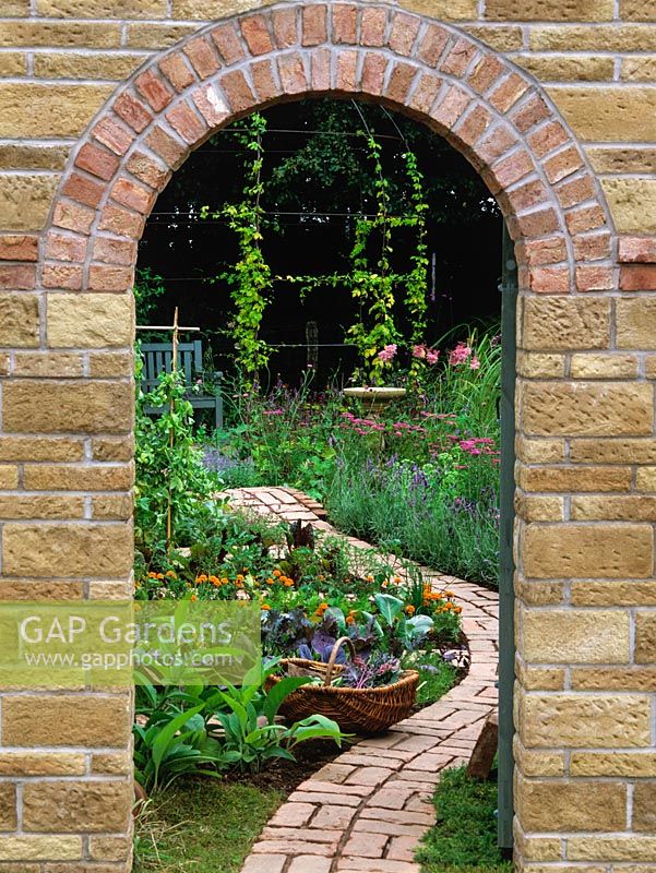 Framed by doorway in brick wall, view of brick garden path winding through vegetable beds to flowers - tagetes, achillea, lavender, sedum, astilbe and hop-clad gazebo.