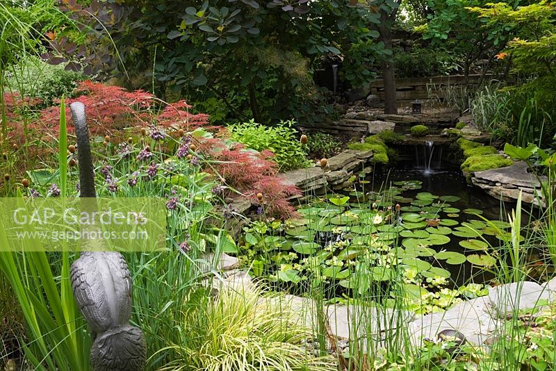 Metal Heron bird sculpture next to purple Tradescantia virginiana - Spiderwort flowers and Acer palmatum inabe-shidare - Japanese Red Maple tree next to pond in backyard garden in summer, Quebec, Canada